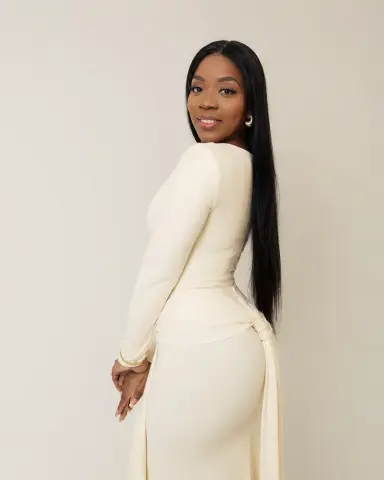 Smiling Black woman in a long-sleeved cream maxi dress posing against a neutral studio background.