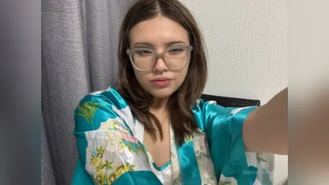 Young woman with glasses wearing a teal floral satin robe in a bright indoor setting.