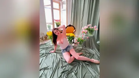 Young woman in a white tank top and denim shorts sitting on green satin fabric surrounded by pink tulips.