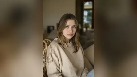 Young woman in a cozy beige sweater sitting in a bright, modern living room with a calm expression.