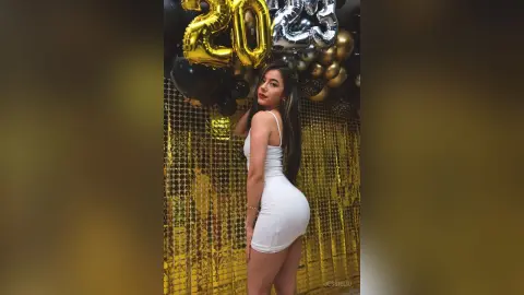 JessieLiu poses in a tight white mini dress against a gold tinsel backdrop with black and gold balloons.