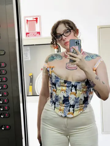 Young woman with tattoos in a floral corset and beige trousers taking a mirror selfie in an elevator.