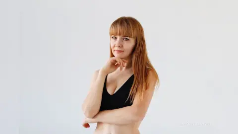 Redheaded woman in a black bikini top posing against a white background with a calm expression.