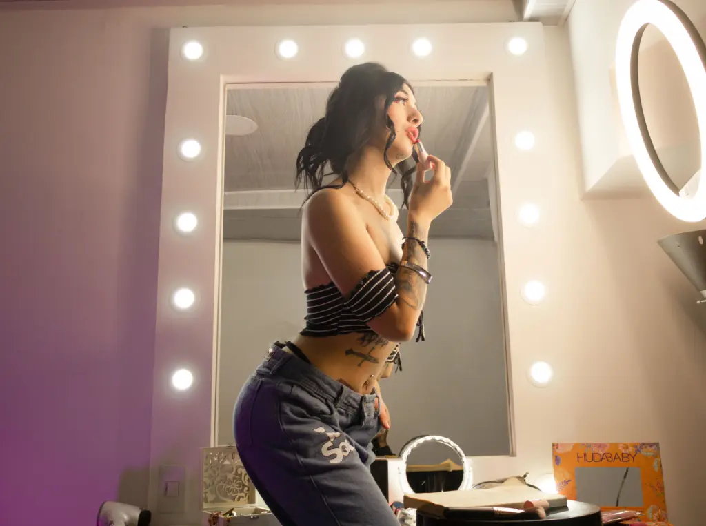 TiffanySantana — Young woman in a black striped crop top and blue jeans posing in front of a lighted vanity mirror.