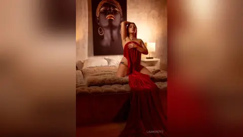 Elegant woman in a flowing red gown posing in a moody, dimly lit bedroom with warm lighting.
