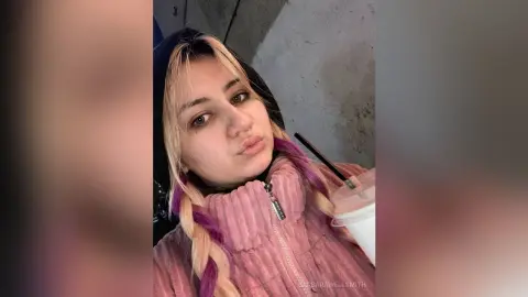 Young woman with blonde and purple braids wearing a pink ribbed jacket while holding a drink in a casual setting.