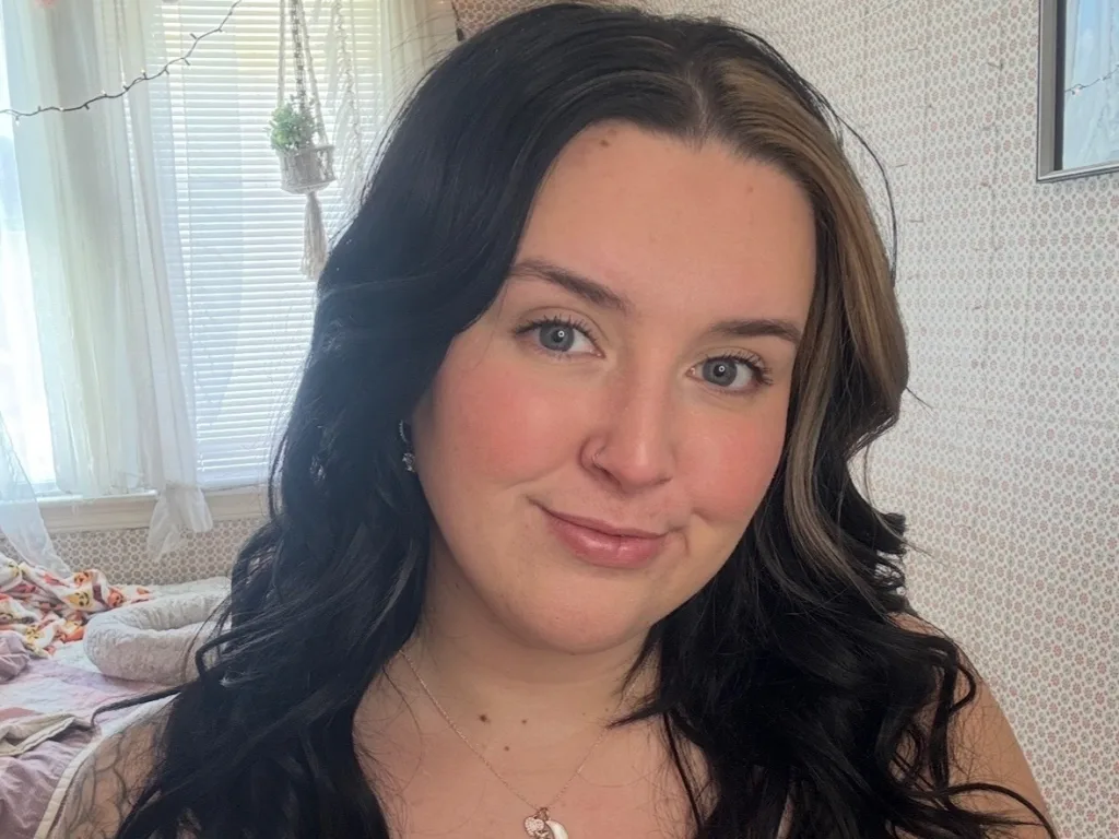 Kassqueen poses with a soft smile, showing her long dark wavy hair and clear skin in a close-up headshot.