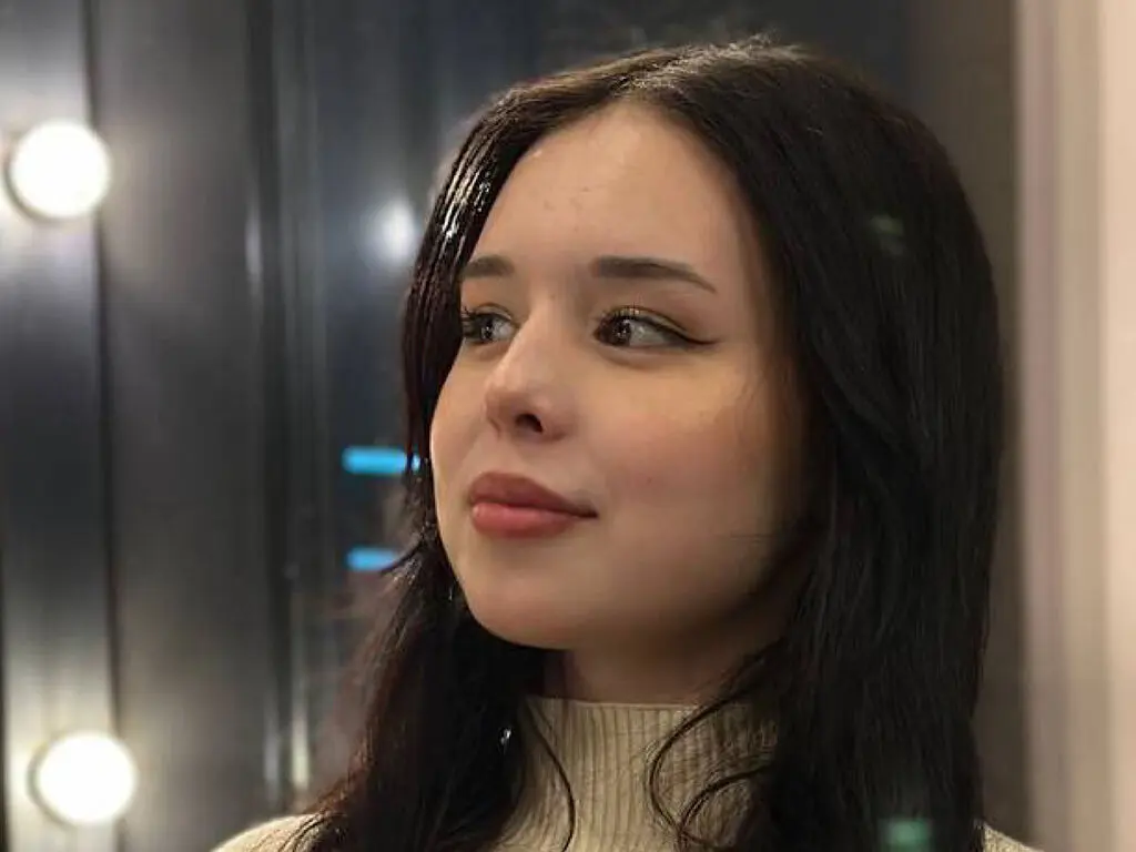 Young woman with dark hair and winged eyeliner wearing a cream turtleneck in a soft, moody indoor setting.