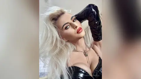 Glamorous blonde woman in a black latex outfit and long gloves posing with a confident, sultry expression.