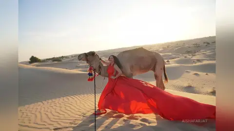 Elegant woman in a flowing red gown posing with a camel in a vast, sunlit desert landscape.