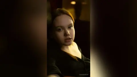 Moody portrait of a young woman in a black top with a nose ring in dim, warm indoor lighting.