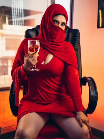 Elegant woman in a red hijab and matching dress holding a glass of wine in a warm, moody setting.