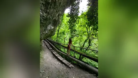 ElizabethSwith stands naked on a wooden walkway in a lush, green jungle setting next to a large rock wall.