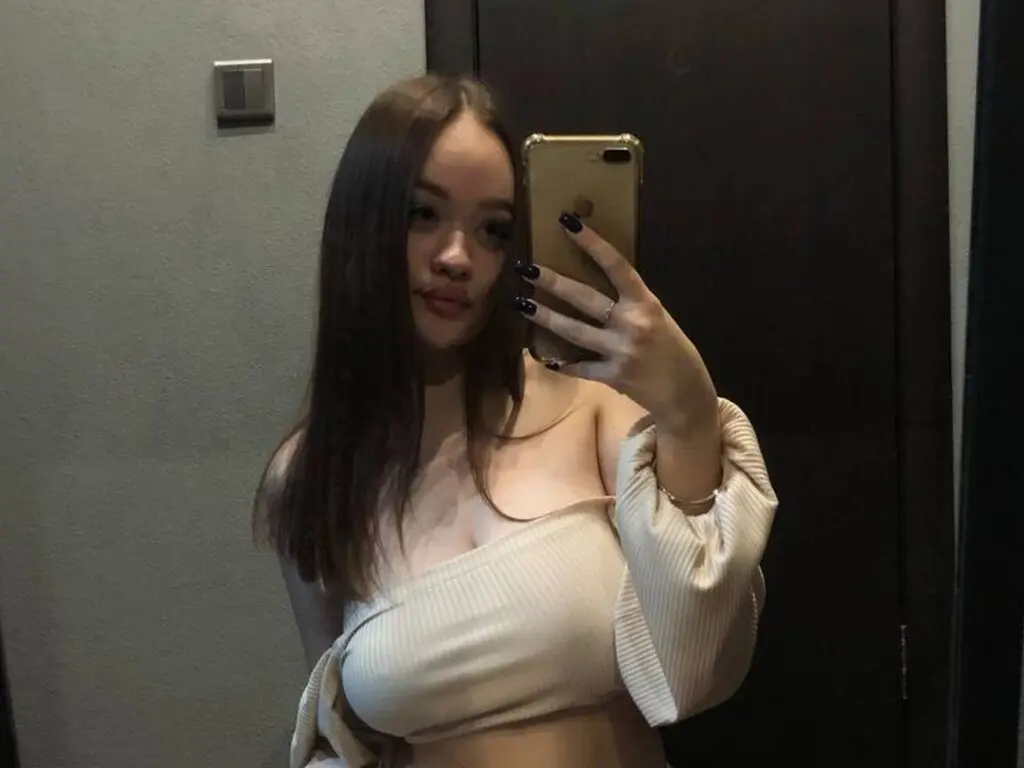 Young woman in a cream ribbed off-the-shoulder top taking a moody mirror selfie in a dimly lit room.