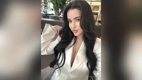 Glamorous woman in a white blazer sitting in a cafe with long wavy dark hair and elegant makeup.