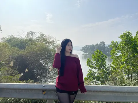 Young woman in a maroon off-the-shoulder sweater posing by a scenic lake under a bright sky.