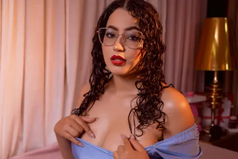 Glamorous woman with curly hair and glasses in a blue off-the-shoulder top posing in a warm bedroom setting.