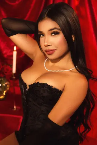 ArianaWoods poses in a black lace corset and pearl necklace against a red velvet background.