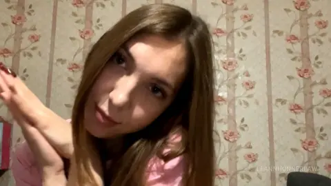 Young woman with long brown hair in a pink shirt smiling against floral patterned wallpaper.