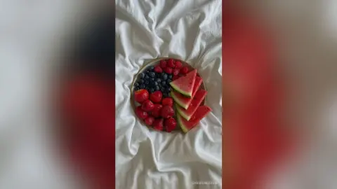 ChristineDavis displays a platter of fresh red strawberries, blueberries, and watermelon slices arranged on a plate over white rumpled bedsheets.