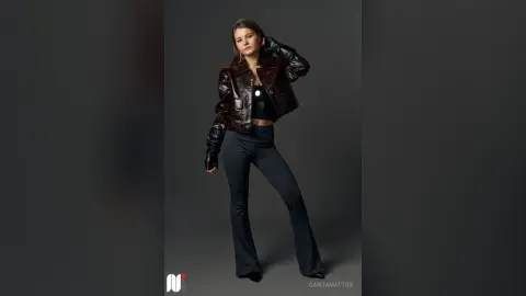 CaritaMattox poses in a dark leather jacket and tight black pants against a gray background.