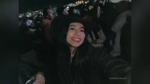 Smiling young woman in a black hat and dark jacket enjoying a lively outdoor night event with a crowd.