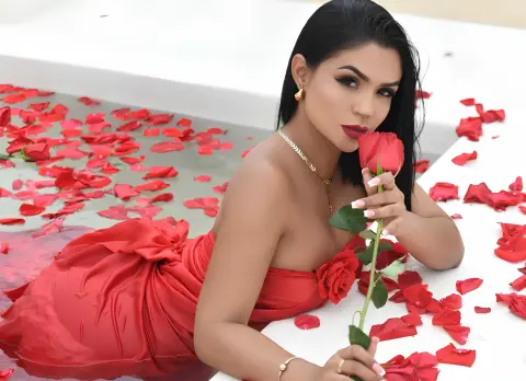 Emilycurtiis with long dark hair wearing a red dress, posing in a white bathtub filled with red rose petals while holding a single rose.