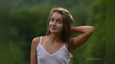 Young woman in a white tank top wearing a flower crown in a lush green meadow with a dreamy, natural mood.