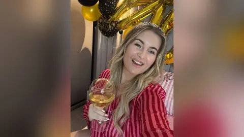 Smiling blonde woman in a pink striped outfit celebrating with a drink and gold balloons at a party.