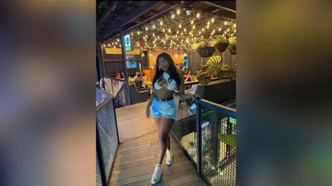 ValeryMei poses in a white bikini top and denim shorts while standing on a wooden balcony under warm hanging lights.