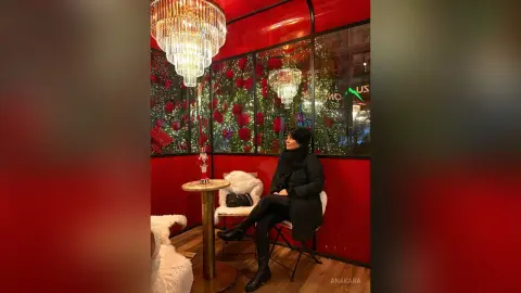 Elegant woman in a black coat sitting in a luxurious red lounge with crystal chandeliers and floral decor.