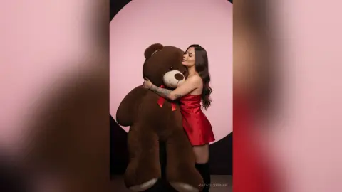 Smiling woman in a red satin dress hugging a giant brown teddy bear against a pink studio background.