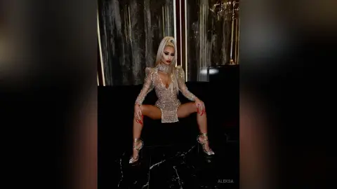 Glamorous blonde woman in a sparkling sequin mini dress sitting on a black sofa in a luxury dark interior.