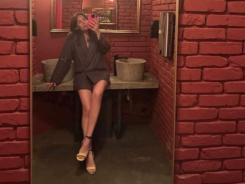 Woman in a chic dark blazer and yellow heels taking a mirror selfie in a rustic red brick bathroom.