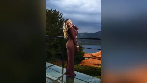 Elegant woman in a long brown evening gown posing on a balcony overlooking a scenic mountain lake at dusk.
