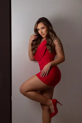EvaSalvatore poses in a tight red mini dress and red high heels against a plain white wall, showing off her long brown hair and shoulder tattoo.