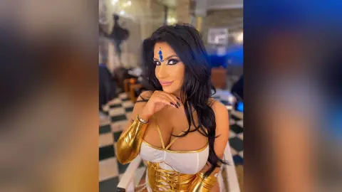 MissyJolie poses with one hand near her chin, wearing a white and gold bustier and long gold gloves with long dark hair.