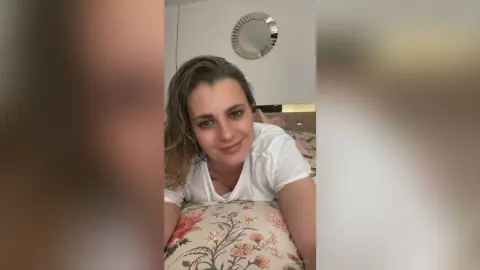 AnishaBee smiles while lying on a floral bed, wearing a white t-shirt with her wet hair falling forward.
