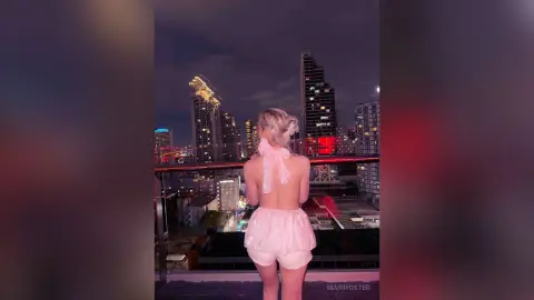 Blonde woman in white satin loungewear overlooking a glowing city skyline at night from a rooftop balcony.