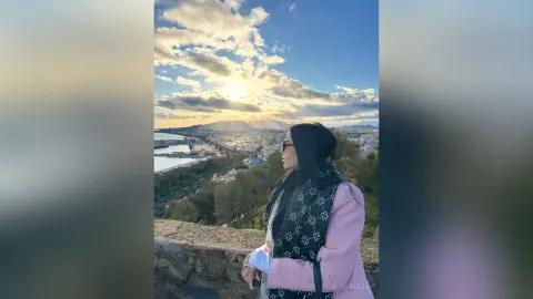 Aleja stands overlooking a coastal city view under a bright, cloudy sky, wearing a black patterned jacket over a white top.