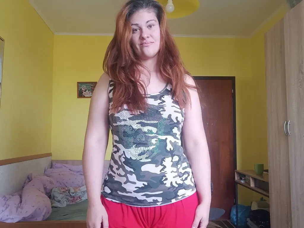MelissaSamantha stands in a yellow room wearing a camouflage tank top and red shorts, letting her long red hair hang down over her shoulders.