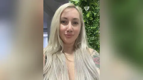 Blonde woman with tattoos posing in a black top against a lush green plant background.