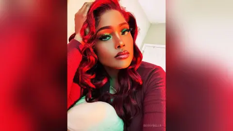 BossyBella — BossyBella poses with her wavy red hair and heavy green eye makeup, wearing a dark long-sleeve top against a plain wall.