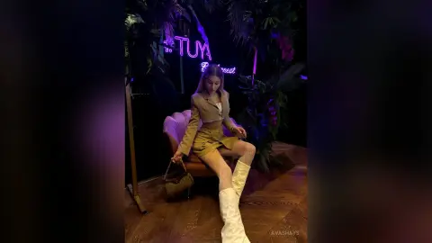Stylish woman in a tan blazer and skirt sitting in a moody neon-lit tropical lounge.