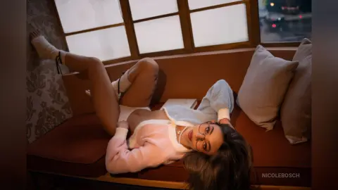 Young woman in a white crop top and shorts relaxing on a brown sofa in a cozy, dimly lit room.