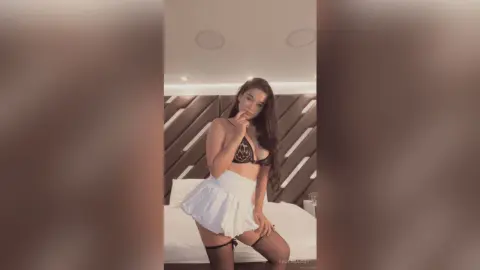Young woman in a white mini skirt and black lace bra posing confidently in a modern bedroom setting.