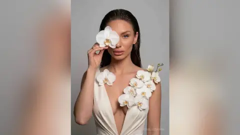 Elegant woman in a white floral gown holding a white orchid against a minimalist grey background.