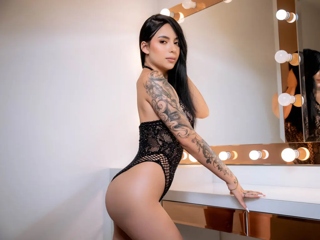 Tattooed woman in a black mesh bodysuit leaning against a lighted vanity mirror in a bright studio.