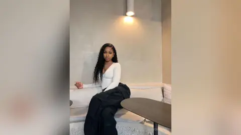 Young woman in a white long-sleeve top and black trousers sitting on a sofa in a modern minimalist room.