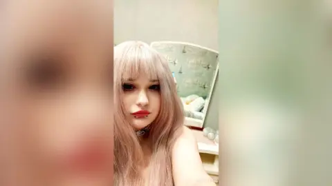EstherTrix poses with her long pink hair and black choker in a bedroom setting.